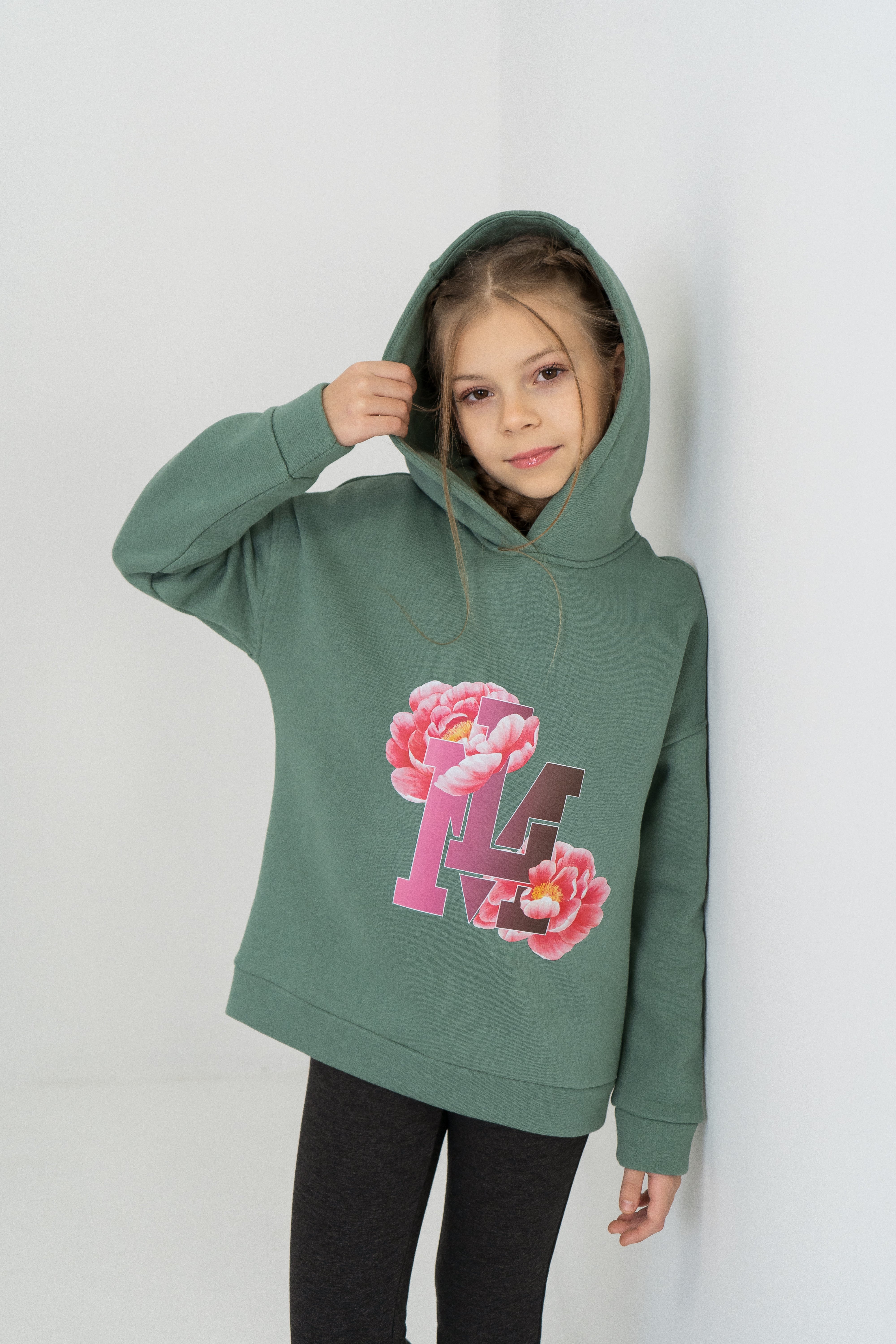 Mädchen zieht die bequeme geräumige Kapuze des grünen Hoodies mit Print von Lemoni Boutique über den Kopf.