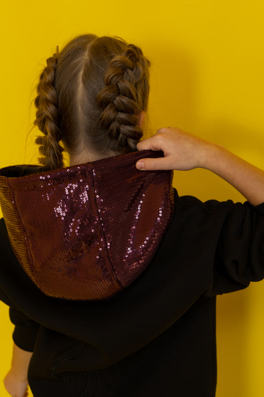 Person wearing a dark hoodie with a shiny, sequined pocket against a yellow background